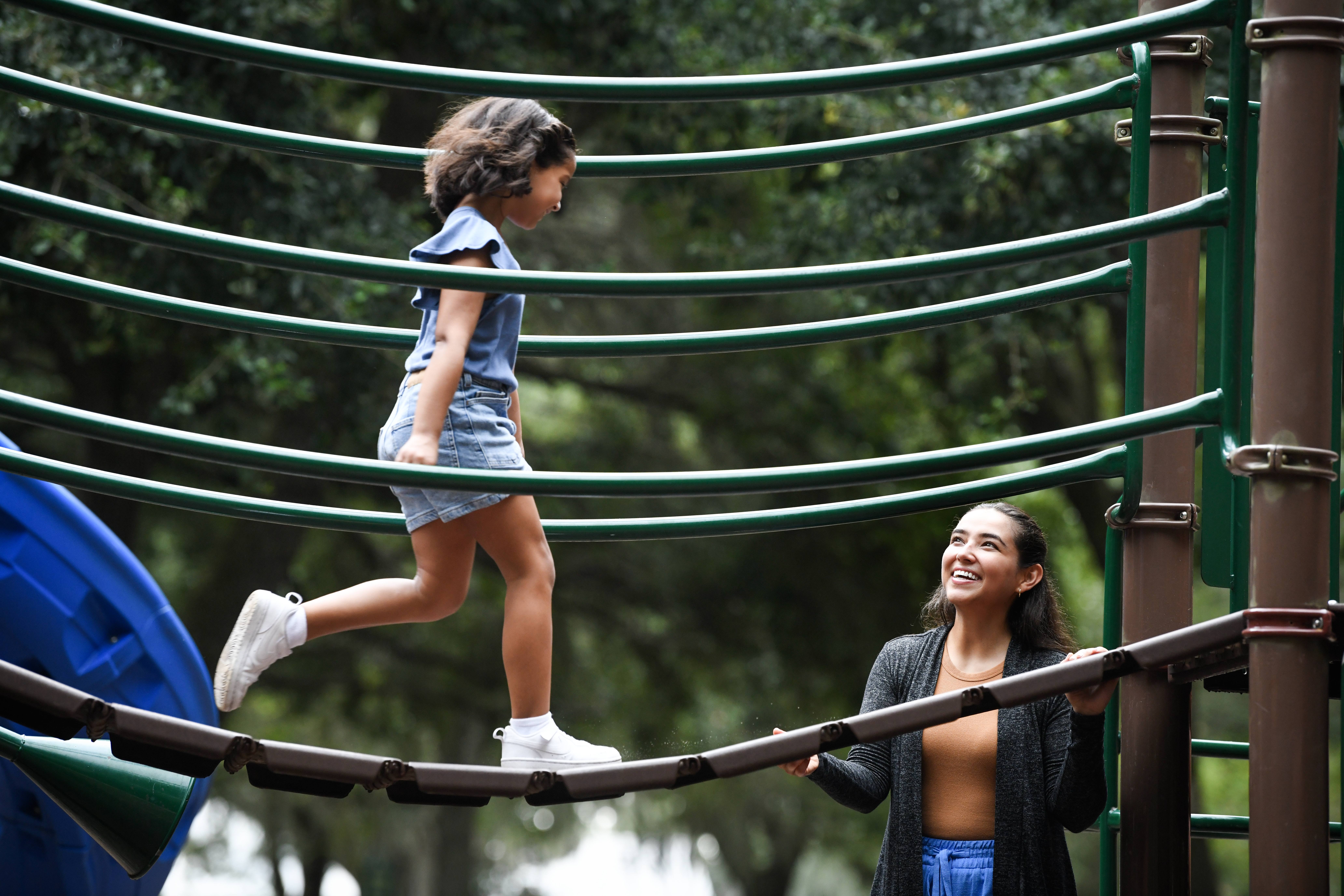 A young child joyfully steps over a playground bridge as a smiling woman watches over her.
